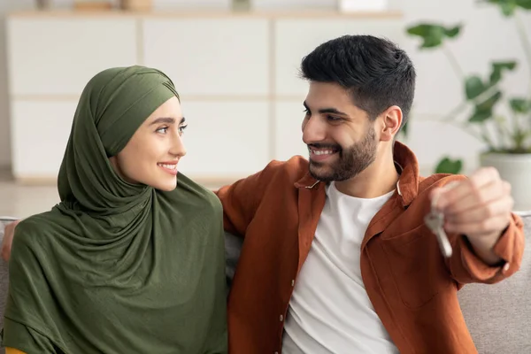 Joyful Middle Eastern Couple Holding New House Key Hugging Sitting On Couch At Home, Smiling To Each Other. Real Estate Purchase And Ownership. Housing For Young Family