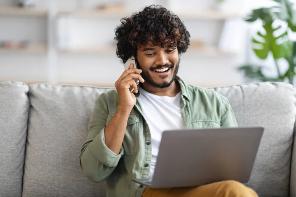 Evde çalışan sakallı, kıvırcık saçlı ve sakallı yakışıklı Hintli adam. Kanepede oturuyor, dizüstü bilgisayar kullanıyor ve iş ortağıyla telefon görüşmesi yapıyor.