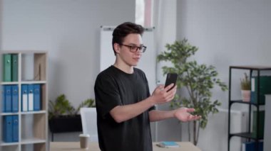 Gözlüklü genç adam akıllı telefon uygulaması üzerinden arkadaşıyla sohbet ediyor, interlokasyonla konuşuyor, ofis içinde duruyor, çekim takibi yapıyor, yavaş çekim yapıyor.