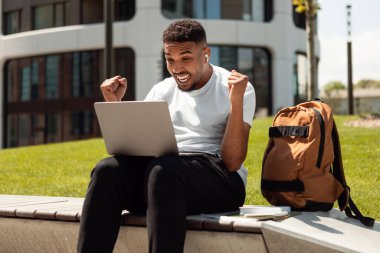 Başarılı bir siyahi adam kutlama yapıyor, yumruklarını sallıyor ve dizüstü bilgisayara bakıyor, şehir merkezinde dışarıda oturuyor. Büyük galibiyet, başarı, inanılmaz online teklif