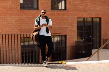 Gözlüklü Afrikalı Amerikalı adam şehirde yürürken akıllı telefon tutuyor ve kameraya bakıp gülümsüyor. Tarz sahibi siyahi adam dışarıda vakit geçiriyor.