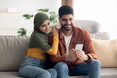 Neşeli Müslüman çift akıllı telefon kullanıyor. Evdeki koltukta oturup internete bakıyorlar. Orta Doğulu eşler cep telefonuyla mesajlaşıyor. Kaynaklar ve İletişim, Harika Uygulama