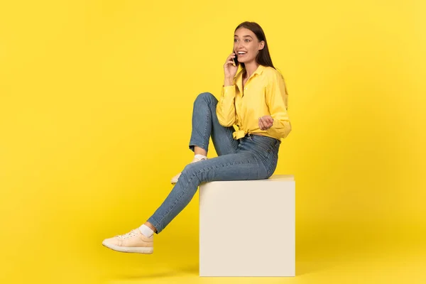 快乐千年女士谈论手机交流坐在立方体上在黄色工作室的背景 有电话交谈的女性呼叫 移动通信 — 图库照片