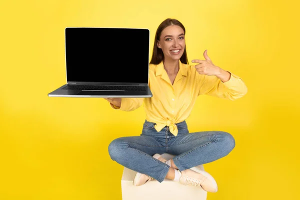 Happy Woman Showing Laptop Computer With Empty Screen Pointing Finger Advertising Great Website Sitting Posing Over Yellow Studio Background, Smiling To Camera. Internet And Technology. Mockup