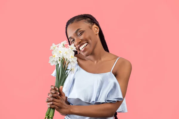 Cheerful millennial black lady holding bouquet of daffodils over pink studio background. Lovely millennial African American woman with bunch of narcissus flowers celebrating holiday