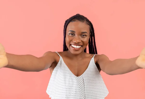 Smiling young black woman taking selfie with smartphone, chatting online on pink studio background. Happy African American lady making self portrait or filming video for social media or her blog