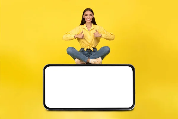 Happy Female Sitting On Huge Cellphone Gesturing Thumbs Up Approving Mobile Offer Smiling To Camera In Studio Over Yellow Background. I Like This Application Concept. Mockup