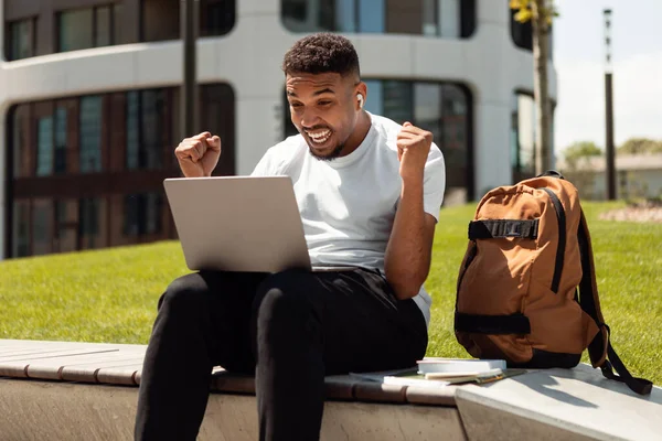 Overjoyed black man celebrating success, shaking fists and looking at laptop computer, sitting outdoors in urban city area. Big win, success, amazing online offer