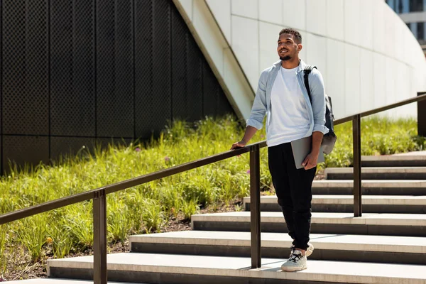 Happy african american man walking in urban city park with laptop and backpack, resting outdoors after work or study, going down the stairs, copy space