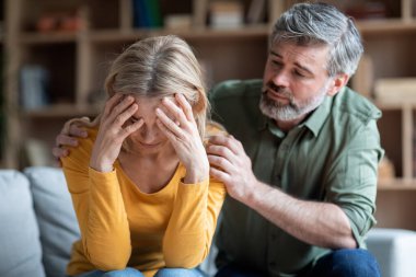 Kadınlardaki orta yaş krizi. Değer veren koca depresif karısını evinde rahatlatıyor, üzgün kadın koltukta oturuyor ve elleriyle yüzünü kapatıyor, depresyon ve ruhsal çöküntü yaşıyor, kapatıyor.