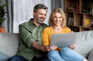 Oturma odasında dizüstü bilgisayar kullanan mutlu orta yaşlı çift, gülümseyen olgun eşler, evde bilgisayarla film seyrediyor ya da internetten alışveriş yapıyorlar, boş alanla yakın çekim yapıyorlar.