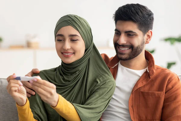 Neşeli Müslüman Eşler Hamilelik Testi Yapıyor Evde Koltukta Oturarak Pozitif Sonuçları Kutluyor. Bebek bekleyen aile çifti. Doğum ve Sağlık Hizmetleri. Seçici Odaklanma