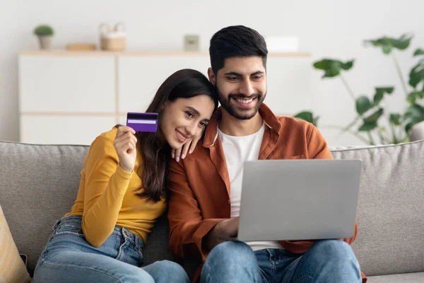 Joyful Middle Eastern Couple Shopping Online Making Payment With Credit Card Using Laptop Computer Sitting On Sofa At Home. Internet Banking And Ecommerce Concept