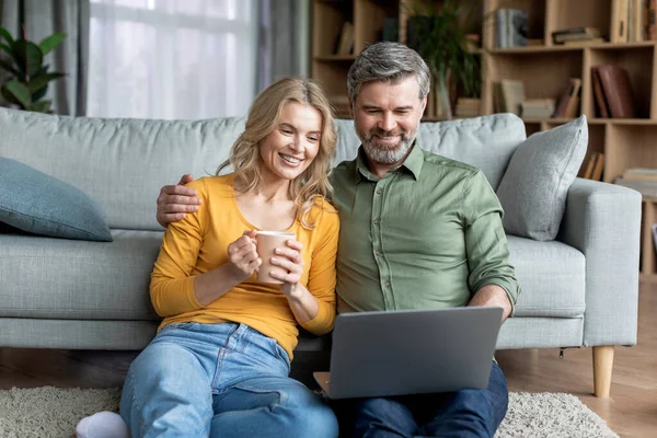 快乐的中年夫妇与笔记本电脑在一起度过时光 微笑着成熟的妻子 丈夫坐在地板上 看电脑屏幕 一起看电影 女人喝咖啡 — 图库照片