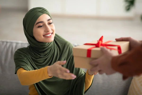 Cheerful Middle Eastern Wife Receiving Gift Box From Husband, Stretching Arms To Wrapped Present Celebrating Family Holiday Sitting On Couch At Home. Gifts Delivery. Cropped, Selective Focus