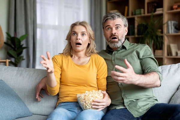 Scary Movie. Shocked Middle Aged Couple Watching Horror Film At Home, Scared Mature Spouses With Popcorn Sitting On Couch In Living Room, Emotionally Reacting To Video Content, Closeup Shot