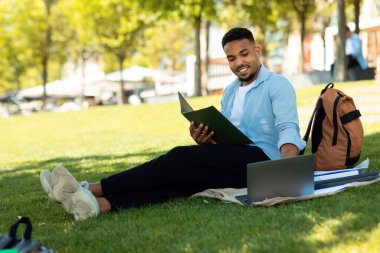 Mutlu siyahi adam deftere notlar yazıyor ve laptopla sınava hazırlanıyor. Şehir parkında oturuyor, boş yer var. Dizüstü bilgisayar ve belge kullanan serbest çalışan
