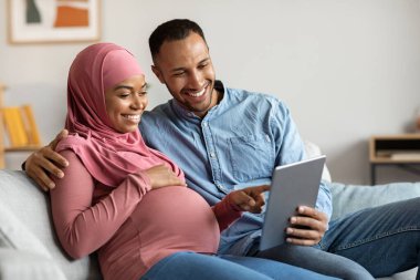 Mutlu siyah hamile çift evde dijital tabletle rahatlıyor, gülümseyen İslami eşler internetten alışveriş yapıyor, bebek için internetten elbise alıyor oturma odasında dinlenirken