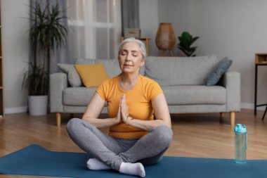 Huzurlu, son sınıf öğrencisi bir kadın, gözleri kapalı meditasyon yapıyor, namaste jestleri yapıyor, yoga minderinde oturuyor. Yoga yapan kadın, iç dengeyi bulan.