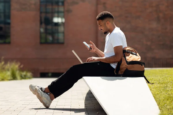 Modern black man with laptop and cellphone sitting outdoors and surfing internet or texting, side view, free space. African american guy using modern laptop and smartphone