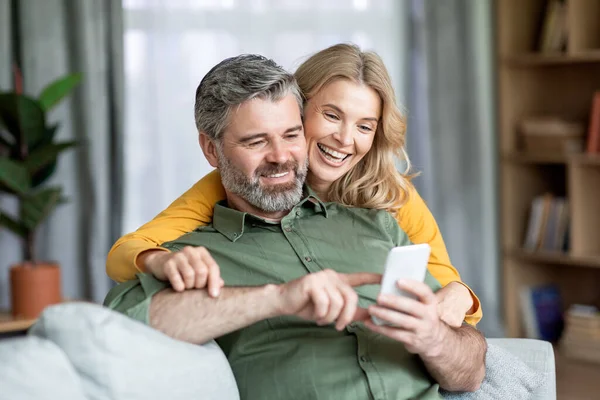 快乐的中年夫妇在家里一起使用手机 上网或上网购物 丈夫在智能手机屏幕上指指点点手指 向他的微笑的妻子展示一些东西 穿上衣服 — 图库照片