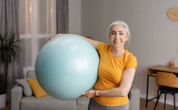 Portrait of positive senior woman holding fitness ball and smiling at camera, exercising in living room at home, leading active lifestyle, keeping fit