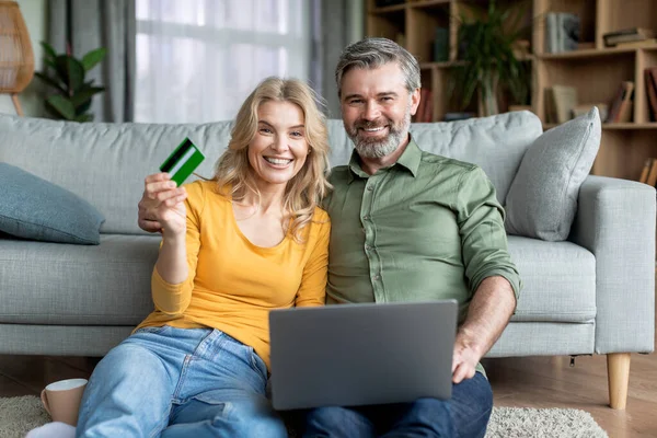 E-ticaret kavramı. Bilgisayarlı ve kredi kartlı mutlu orta yaşlı çift evden alışveriş yapıyor, olgun eşler oturma odasında bilgisayar başında oturuyor ve kameraya gülümsüyor,