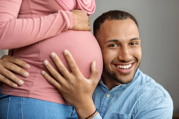 Happy black man embracing belly of his pregnant muslim wife in hijab, excited husband hugging spouse tummy and listening baby kicks, enjoying upcoming fatherhood, closeup shot, cropped image