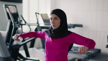 Müslüman bayanlar için spor eğitimi. Genç aktif ortadoğulu kadın spor salonunda egzersiz yapıyor, dambıllarla pratik yapıyor, ağır çekimde.