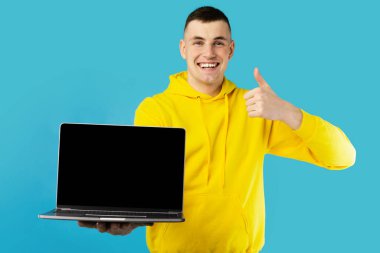 Neşeli Serbest Çalışan Adam, boş ekranlı dizüstü bilgisayar gösteriyor, başparmak hareketi yapıyor, harika bir web sitesi, Mavi Stüdyo 'nun arkasında duruyor, kameraya gülümsüyor. İnternet Teknolojisi