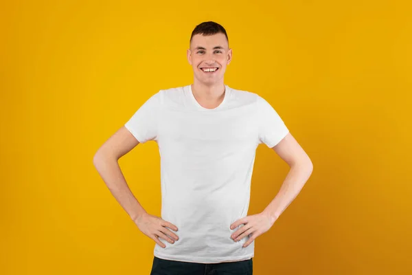 快乐的千禧年男人牵着小腿微笑着站在黄色背景的工作室里 穿着白色T恤的正人君子中枪 男性时装概念 — 图库照片