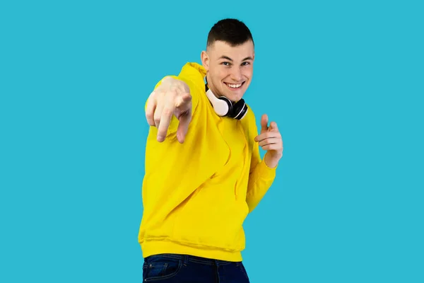 I Choose You. Happy Millennial Man With Wireless Headphones Pointing Finger At Camera Smiling Posing Over Blue Studio Background, Wearing Yellow Hoodie. Youre Next Concept