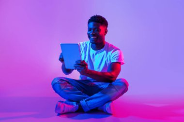 Tablet bilgisayar kullanan yakışıklı siyahi adam, çapraz bacaklı neon ışıkta oturuyor. Neşeli Afrikalı Amerikalı adam internette çalışıyor ya da okuyor, internette video konferansı yapıyor.
