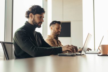 Bilgisayarlar kullanarak odaklanmış şirket çalışanları, modern açık uzay ofisinde bilgisayar aygıtlarıyla çalışıyorlar. Erkek iş arkadaşları iş projesini geliştiriyor, birlikte çalışıyorlar.