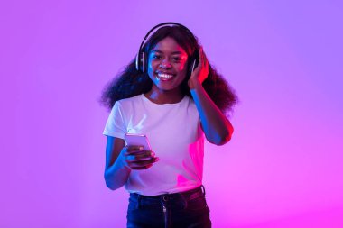 Kablosuz kulaklıklı, neon ışıkta akıllı telefon kullanan mutlu bin yıllık siyahi kadın. Güzel genç Afrikalı Amerikalı kadın yeni mobil müzik uygulamasını kontrol ediyor, internetten sesli kitap dinliyor.