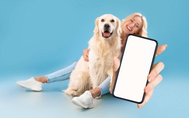 Güzel genç bir kadın köpeğiyle oturuyor ve kamerada beyaz ekranlı büyük boş akıllı telefonu gösteriyor. Mutlu bir evcil hayvan sahibi mavi arka planda poz verirken yeni bir mobil uygulama veya web sitesi öneriyor.
