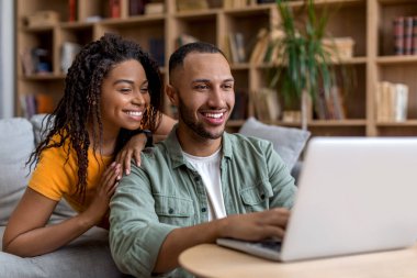 Mutlu Afro-Amerikan eşleri kanepede dizüstü bilgisayarlarıyla oturuyor, evde birlikte eğleniyor, internetten yeni mobilyalar arıyor, web sörfü yapıyor, kopyalama alanı arıyor.