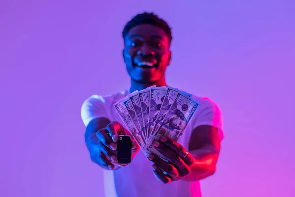 Portrait of excited young black man showing new car key and money in neon light, selective focus. Millennial African American guy buying or winning automobile. Auto loan concept