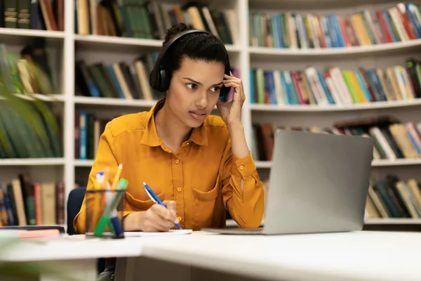 ノートパソコンを見て オンラインで学び ノートを取り 図書館の机に座っている女性学生に焦点を当てています 遠隔講義 遠隔研究 現代教育の概念 — ストック写真