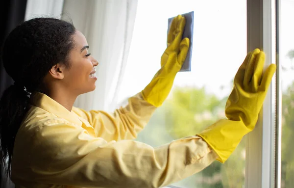 Cheerful Black Female Cleaning Window With A Rag And Detergent Wearing Yellow Rubber Gloves Doing Housework Standing At Home. Maid Service Concept. Side View Shot