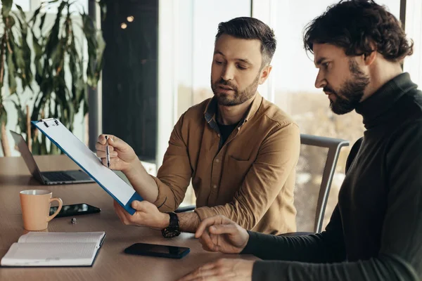 Erkek girişimciler iş belgelerini inceliyor, anlaşma ya da sözleşme üzerinde çalışıyor, modern ofiste masa başında oturuyor. İş adamları anlaşmayı tartışıyor, işbirliği yapıyorlar.