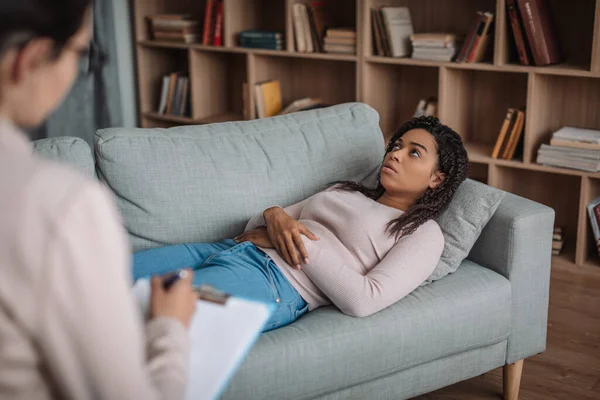 Serious african american millennial woman lies on sofa at consultation with doctor in office clinic interior. Psychological therapy, mental problems and treatment, medicine, support, professional help