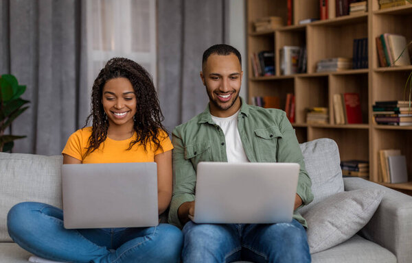 Young african american guy and his girlfriend using modern laptops and smiling while sitting on couch at home. Young black couple working online via pc computers