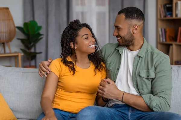 Romantik Afrikalı Amerikalı eşler evdeki kanepede dinleniyorlar, konuşuyorlar, kucaklaşıyorlar ve birlikte vakit geçiriyorlar, oturma odasında dinleniyorlar. Mutlu çift birbirine bakıyor ve gülümsüyor.