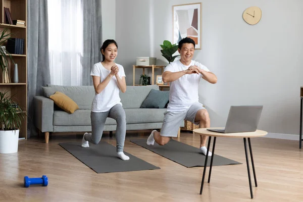 Strong chinese spouses exercising together at home, watching fitness video, smiling asian man and woman in sportswear standing on yoga mats in warrior position, having online yoga class, copy space