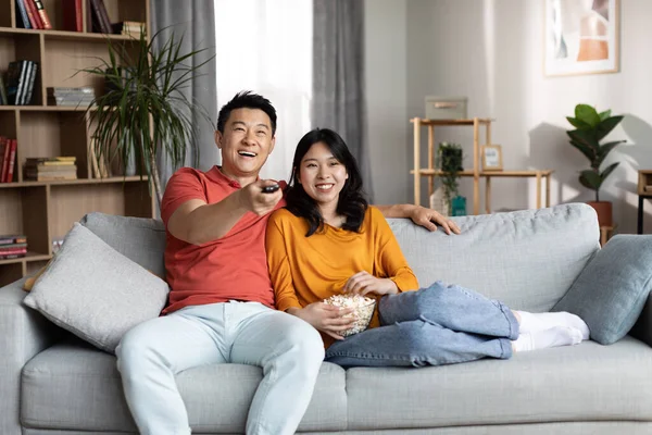 Watching comedy film or TV series. Loving chinese couple resting on sofa, spending weekend at home together, holding remote controller, eating popcorn and laughing, copy space