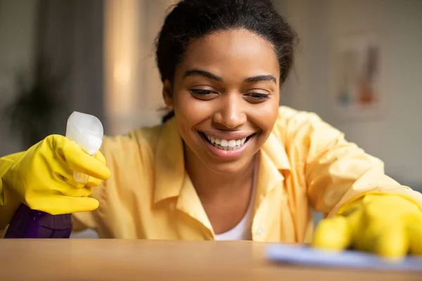 Happy African American Woman Dusting Shelf With Rag And Detergent Spray Caring For Wooden Furniture Cleaning At Home, Wearing Yellow Gloves. House Chores. Selective Focus