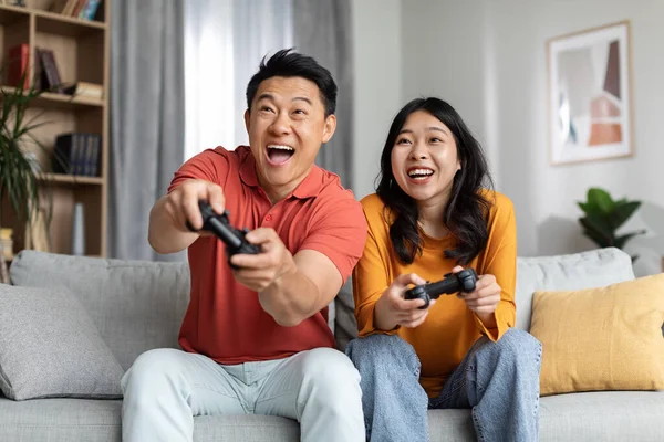 兴奋的韩国夫妇在家里玩电子游戏 快乐的亚洲男人和女人坐在沙发上 手握操纵杆 玩得开心 周末愉快 复制空间 — 图库照片
