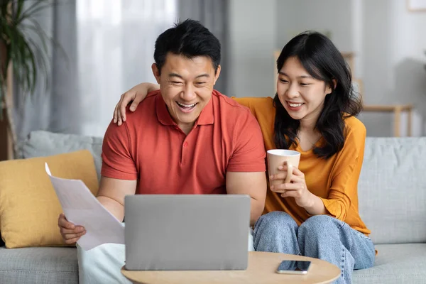 Smiling asian spouses paying bills online while staying home, sitting on couch, using laptop, happy chinese man holding papers and banking on Internet, cheerful woman drinking tea, copy space