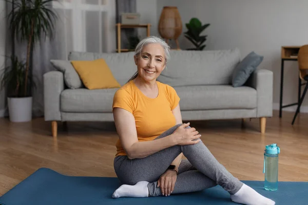 Healthy lifestyle concept. Happy senior woman sitting on sports yoga mat at home in living room and smiling at camera, copy space. Elderly female doing rehab exercises